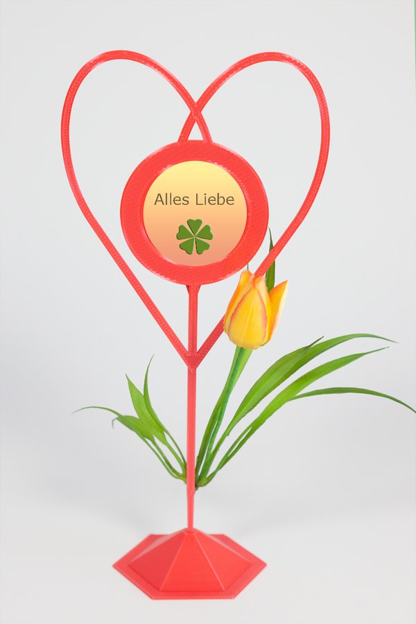 Beispiel-Dekoration - Blumenstecker "Herz hellrot"  mit Standfuß - an dem Standfuß befestigt ist eine gelbrote Tulpenblüte mit einigen Blättern - eingesetztes Foto: gelb-roter Hintergrund mit grüner Kleeblatt-Grafik und Text "Alles Liebe"